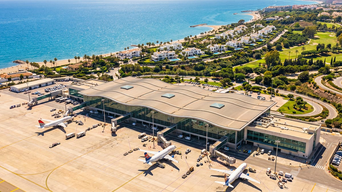 Aerial view of Málaga Airport in Costa del Sol, showcasing its modern terminal, vibrant Mediterranean coastline, luxury villas, and palm trees under clear blue skies.